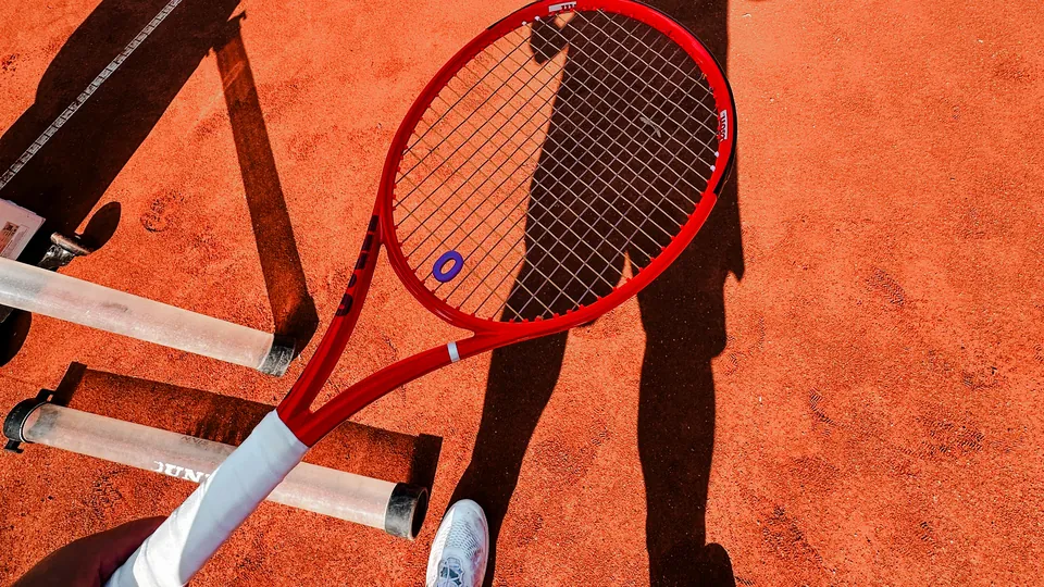 Schatten eines Tennisspielers auf dem Sandplatz, im Vordergrund ein roter Tennisschläger
