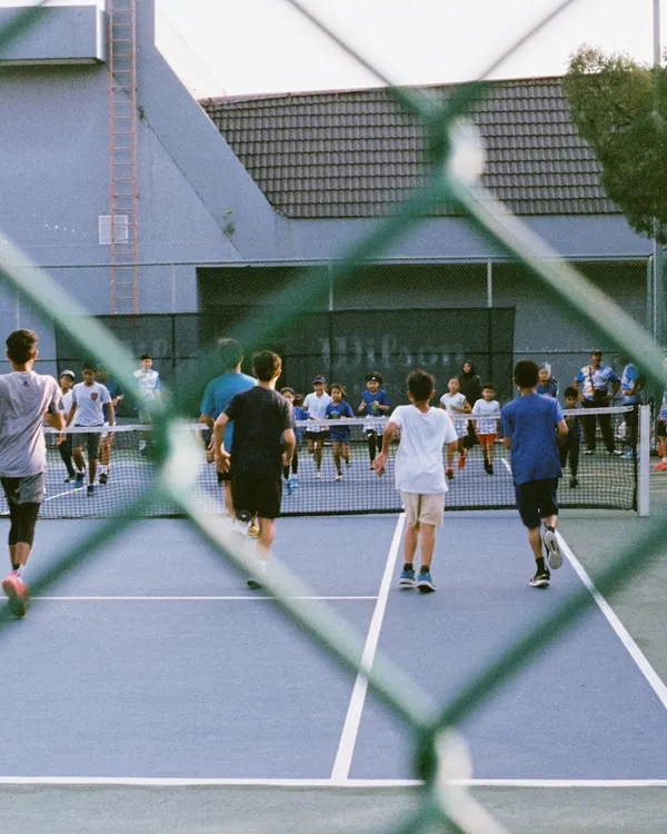 Viele Spielerinnen und Spieler auf einem Tennisplatz, durch den Zaun fotografiert